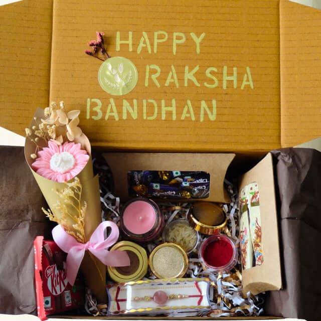 Gift box with 'Happy Raksha Bandhan' text, flowers, and small items on a brown background