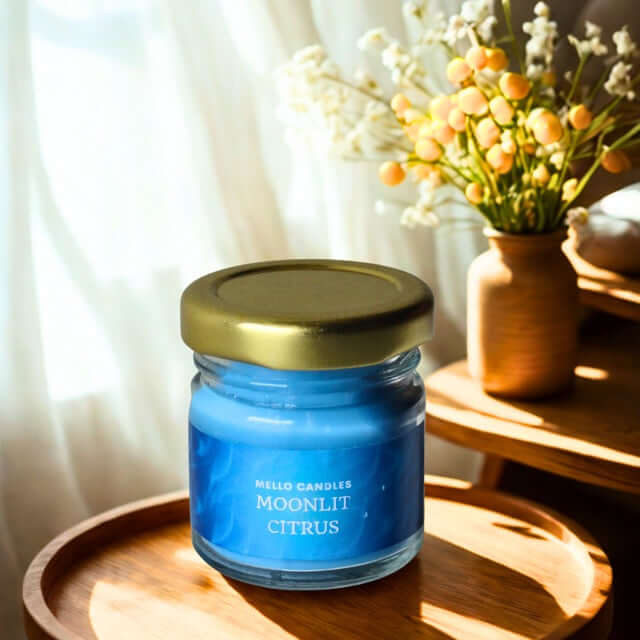 Moonlit citrus candle in a glass jar with a gold lid on a wooden surface, with a vase of flowers in the background.