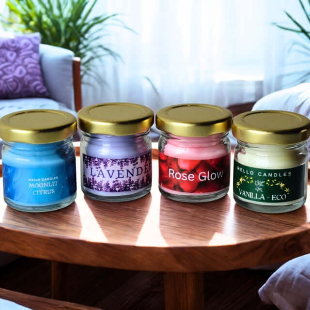 Four candles with gold lids on a wooden table, labeled 'Moonlit Citrus', 'Lavender', 'Rose Glow', and 'Vanilla Eco'.