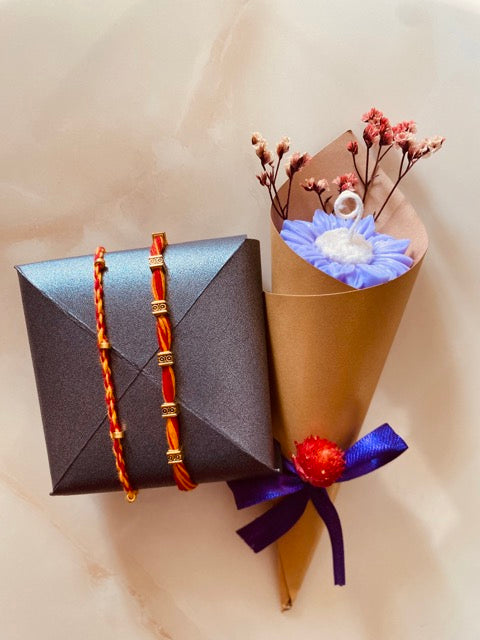 Gray gift box with a red and gold rakhi, a brown paper cone with candle flowers